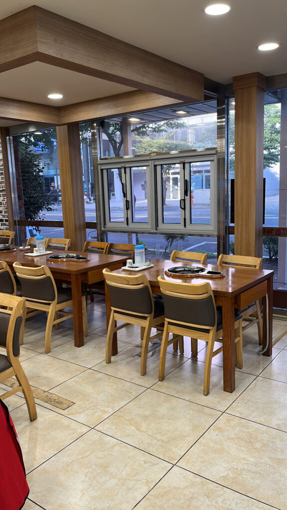 Bright main dining hall featuring large glass windows and wooden tables equipped with built-in gas stoves.
