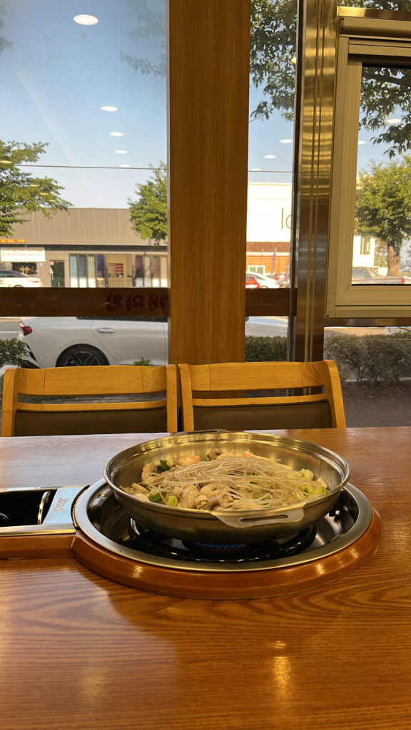 A pot of uncooked Nakgopsae placed on a table stove by a large window with an outside street view.