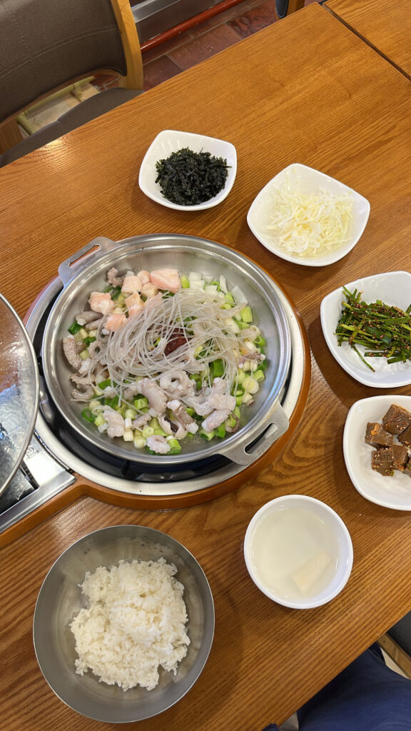 A table set with raw Nakgopsae ingredients in a pot, a bowl of rice, cold radish soup, and side dishes.