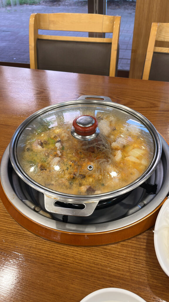 A stainless steel pot covered with a glass lid, with Nakgopsae boiling over a gas stove.
