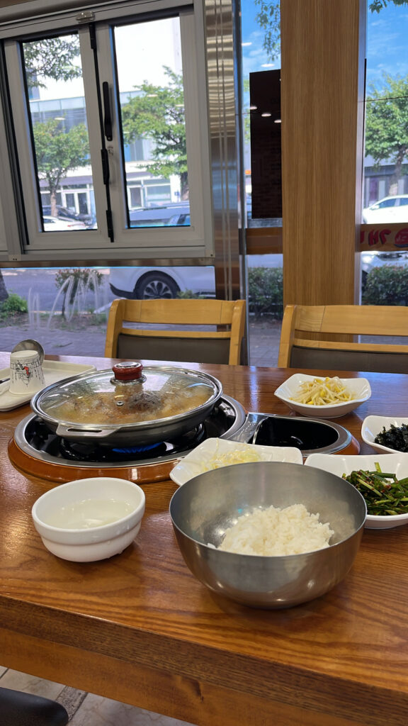 A dining table with boiling Nakgopsae, side dishes including bean sprouts and chives, and a bowl of rice.