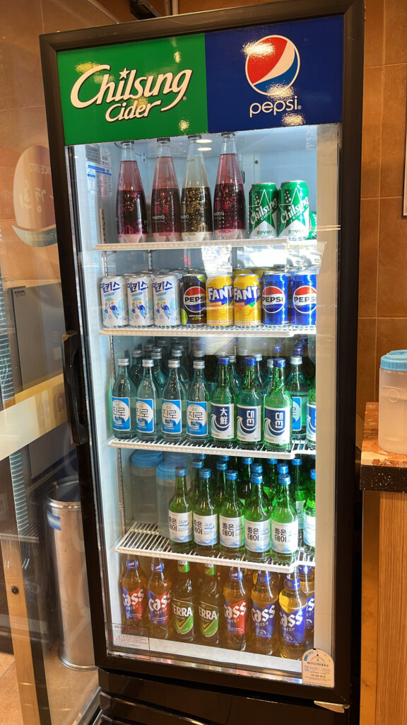 A commercial drink refrigerator filled with various Korean beverages, soft drinks, soju, and beer.