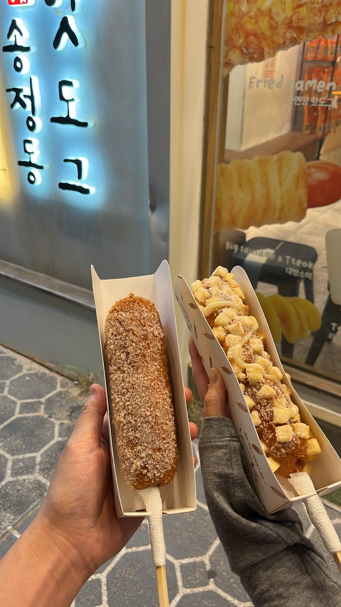 Holding two different crispy corn dogs in front of the store at night